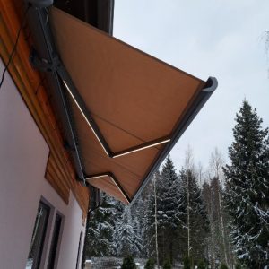 brun retractable-markis taloverannalla talvisessa maisemassa, taustalla lumisia kuusia